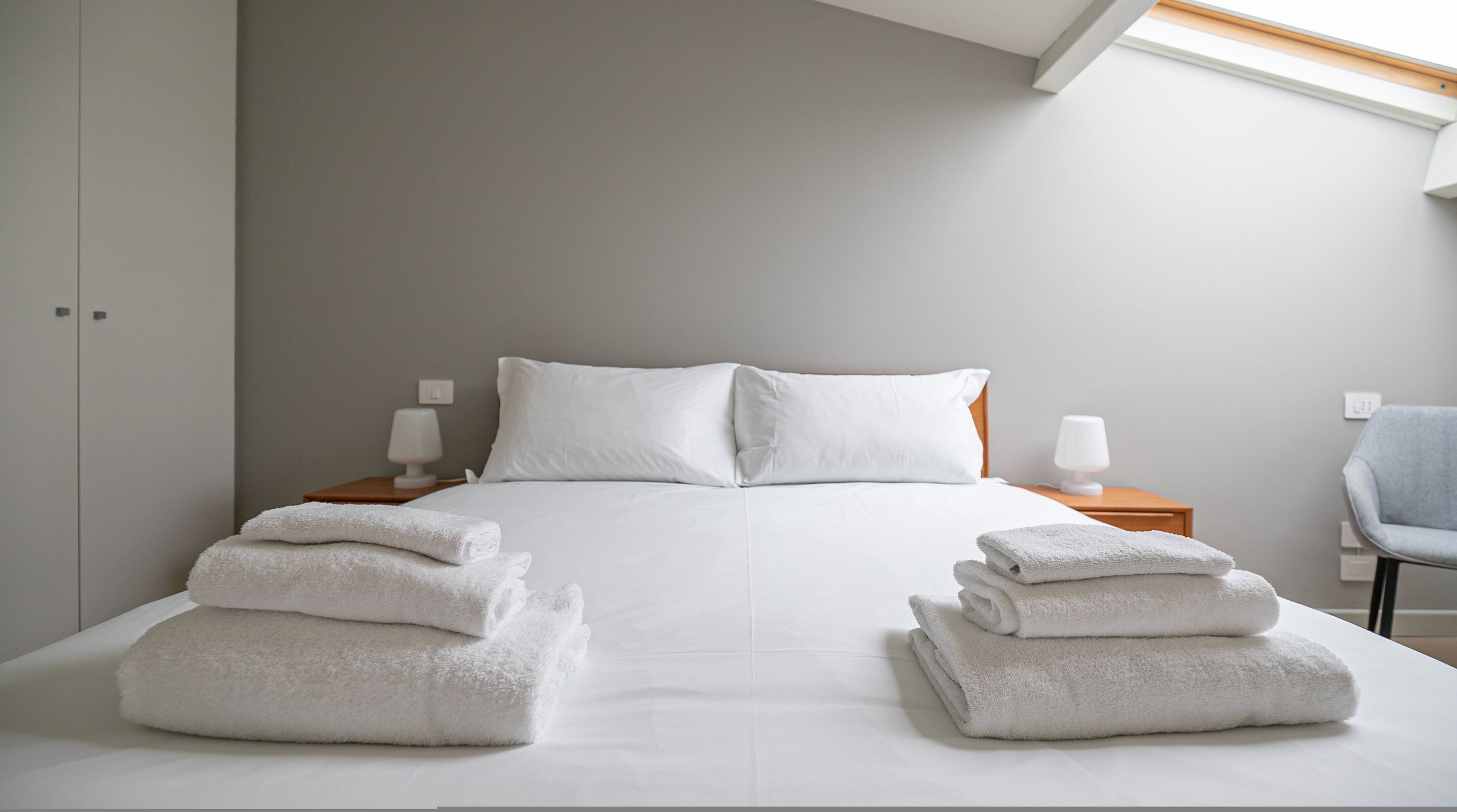 A neatly made bed with white linens and two stacks of folded towels at the foot of the bed. There are two bedside tables with lamps, a grey chair, and a wardrobe in the background.