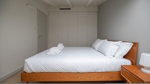 Een net opgemaakt bed met witte lakens en kussens, een houten hoofdbord en een nachtkastje met een witte lamp.