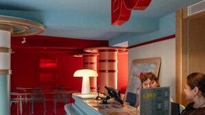 A reception desk with a blue counter and a red sign above it. There are chairs and a table in the background. - The Boc Hostels Beach (Palma de Mallorca)