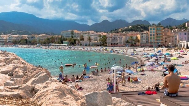 A crowded beach with people sunbathing and swimming, surrounded by buildings and mountains in the background.