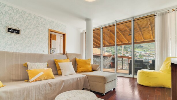 A modern living room with a sofa, yellow cushions, a round table, and a view of a balcony with a wooden roof. - Alfa I (Machico)
