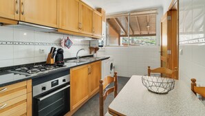 A kitchen with wooden cabinets, a stove, a washing machine, and a dining area with a table and chairs. - Alfa I (Machico)