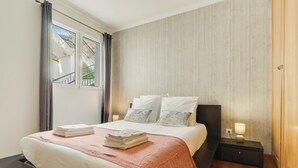 A neatly made bed with a pink blanket and folded towels, a bedside table with a lamp, and a window with curtains. - Alfa I (Machico)