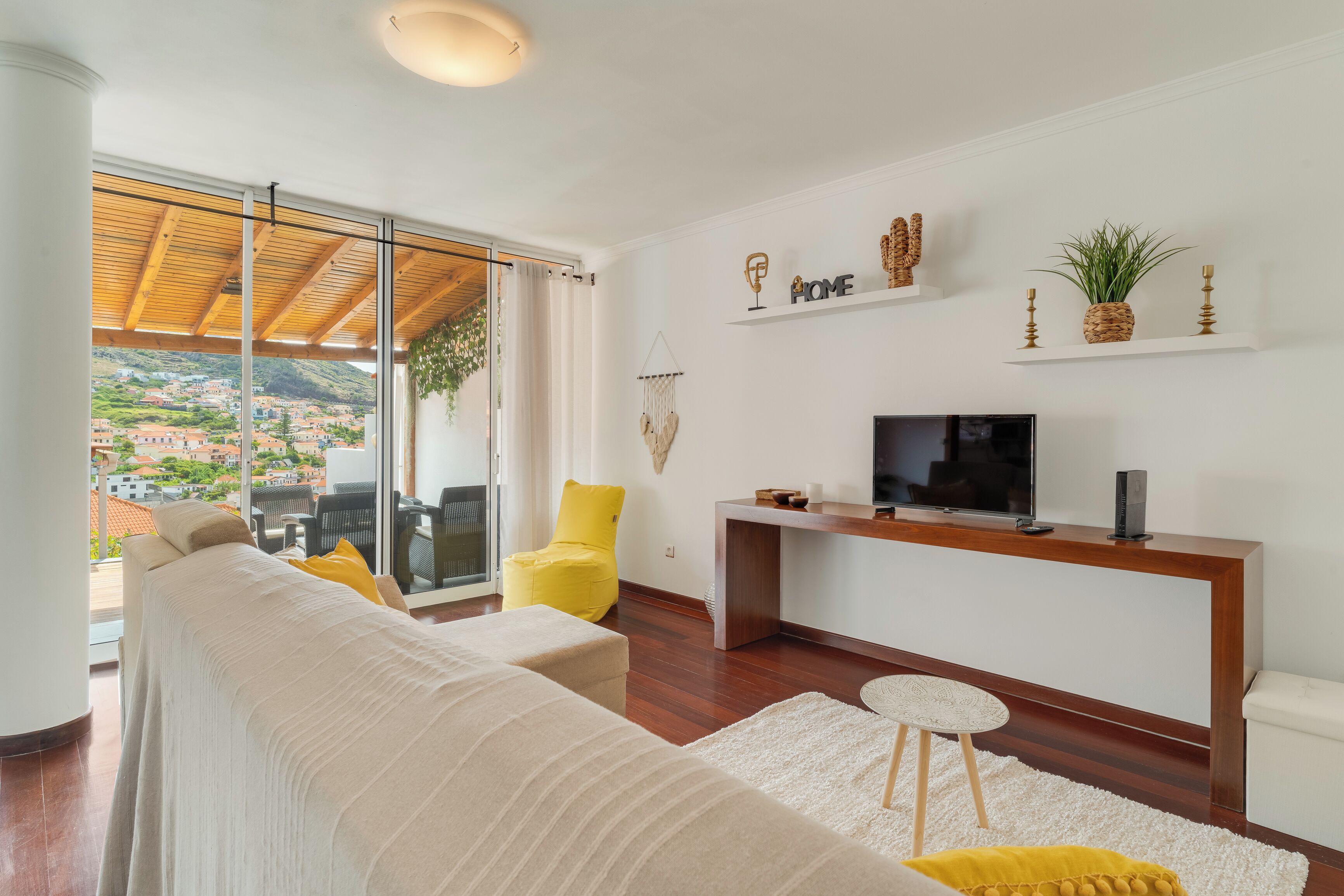 Een moderne woonkamer met een bank, gele fauteuil, tv en uitzicht op een heuvelstad.