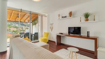 Een moderne woonkamer met een bank, gele fauteuil, tv en uitzicht op een heuvelstad.