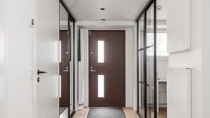 Een gang met een bruine deur, witte muren en een zwarte mat die naar een kamer met een raam leidt.
