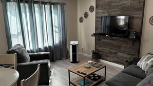 Un salon moderne avec un téléviseur à écran plat fixé à un mur en bois, des canapés gris, une table basse avec des télécommandes, une table à manger et une grande fenêtre ornée de rideaux légers.