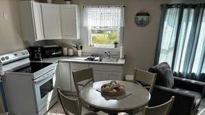 Une cuisine compacte avec des meubles blancs, un four à micro-ondes, une machine à café et un évier. On y trouve une table à manger avec des chaises et une horloge au mur.