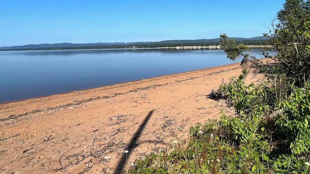 Une plage de sable bordée d’un lac paisible, entourée de verdure et de collines lointaines.