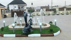A fountain with multiple water jets in a circular arrangement, surrounded by a grassy area and a low wall. - G- throne Hotel and Suites (Port Harcourt)