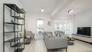 Una sala moderna con un sofá gris, una estantería negra, un televisor y un comedor con sillas y una mesa.