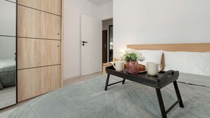 Un dormitorio con cama, mesita de noche con bandeja que sostiene una taza, florero con flores y armario de madera.