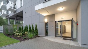 Una entrada de edificio moderna con puerta de vidrio, un paseo empedrado y una zona ajardinada con arbustos y un árbol pequeño.