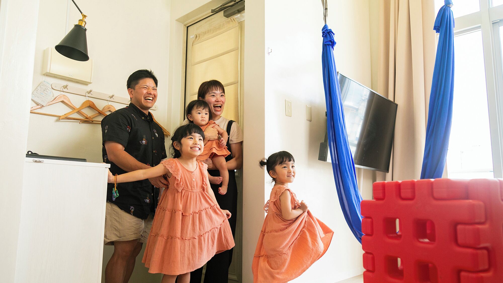 Una familia en una habitación con televisión, una puerta y un ventanal con cortinas.