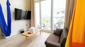 Una sala moderna con sofá, televisor montado en la pared, mesa de madera con control remoto y un balcón con mesa y sillas.