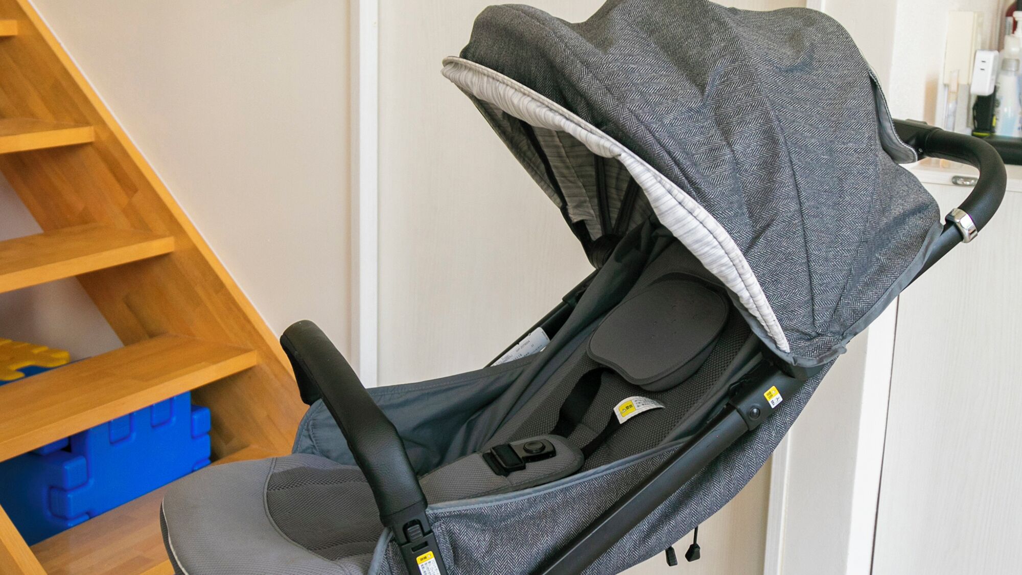 Een kinderwagen met een grijs herringbone patroon, een zwarte handgreep en een grijze zitting.