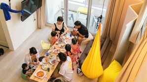 Una familia cenando en un cuarto moderno y luminoso, con un ventanal grande, un sofá y un sillón colgante.