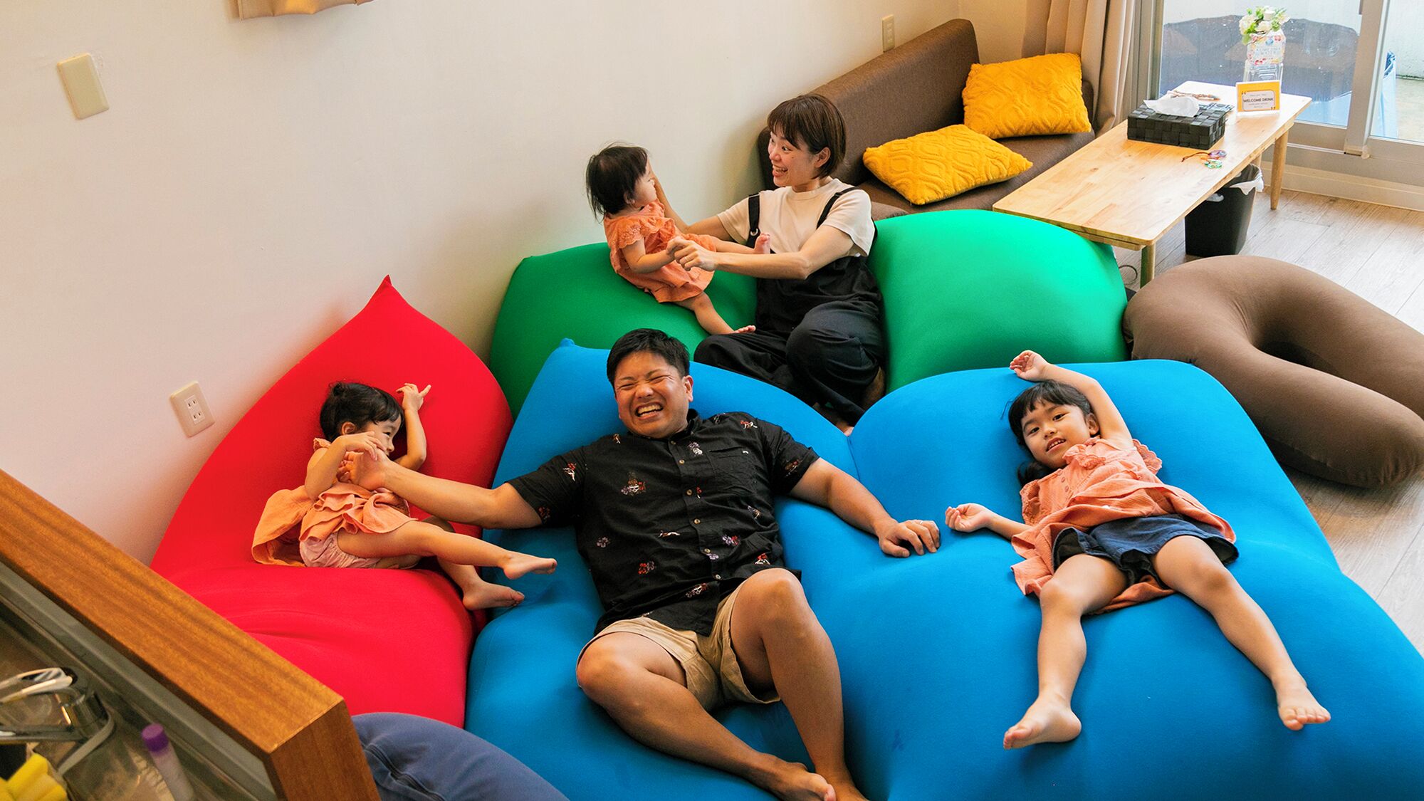 Une famille de quatre personnes est assise sur des canapés modulaires aux couleurs vives, dans un salon moderne et lumineux, avec une table en bois et une fenêtre en arrière-plan.