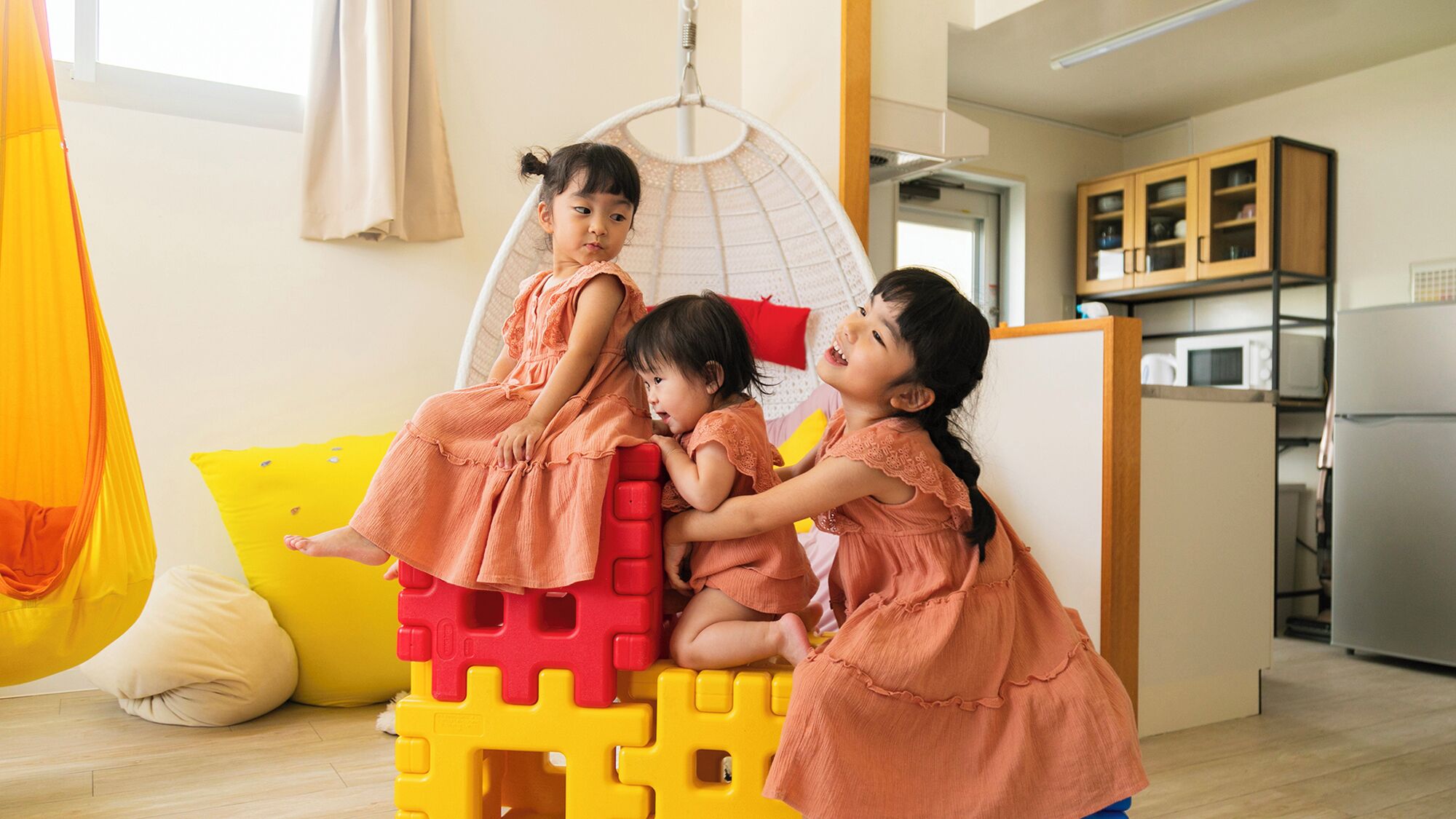 A play area with a hanging chair, a yellow and red play structure, and a kitchen in the background.