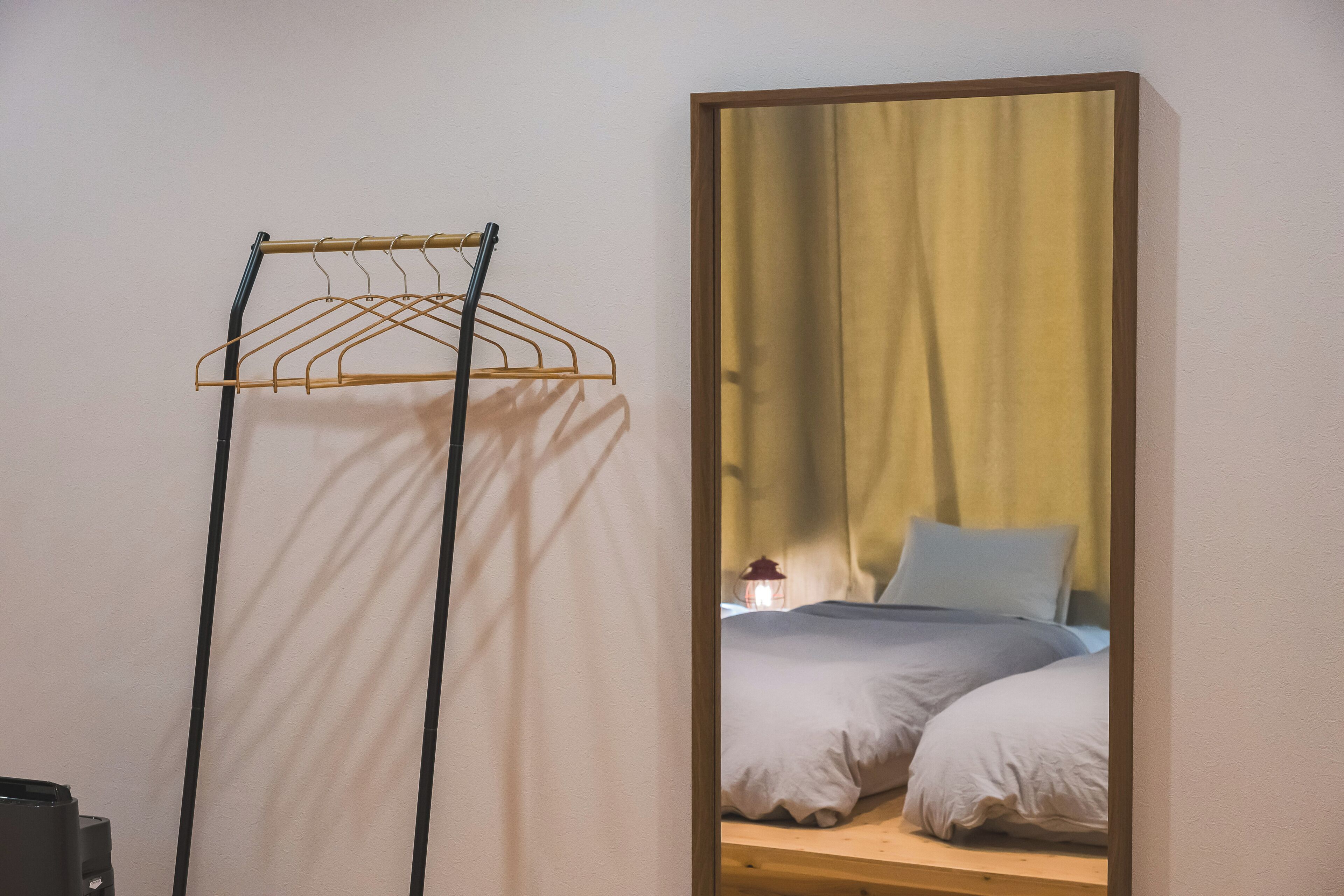 A room with a bed, a hanging clothes rack, and a mirror reflecting a curtain and a lamp.