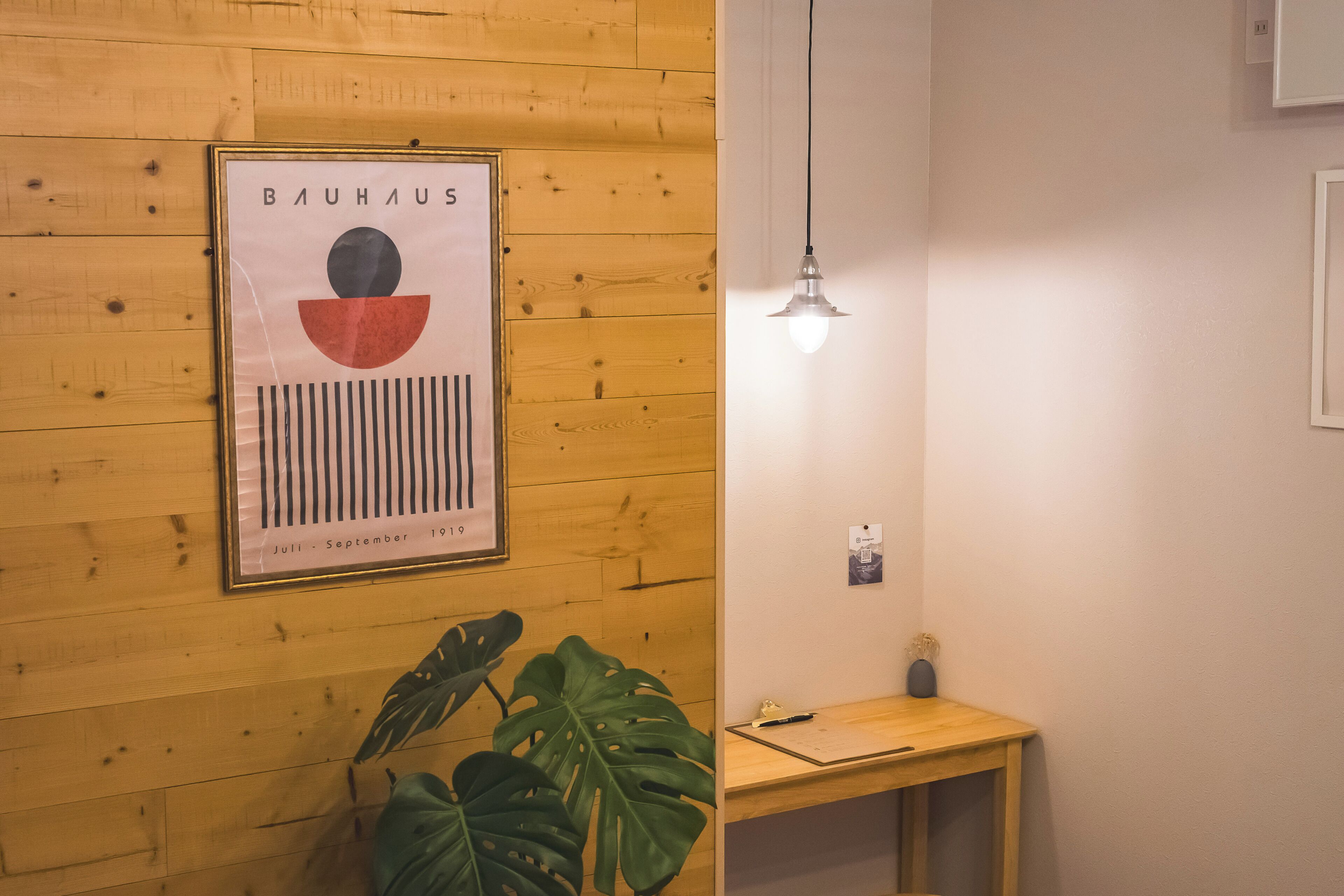 Una pared de madera con un cartel Bauhaus enmarcado, una planta en maceta, un escritorio y una lámpara colgante.