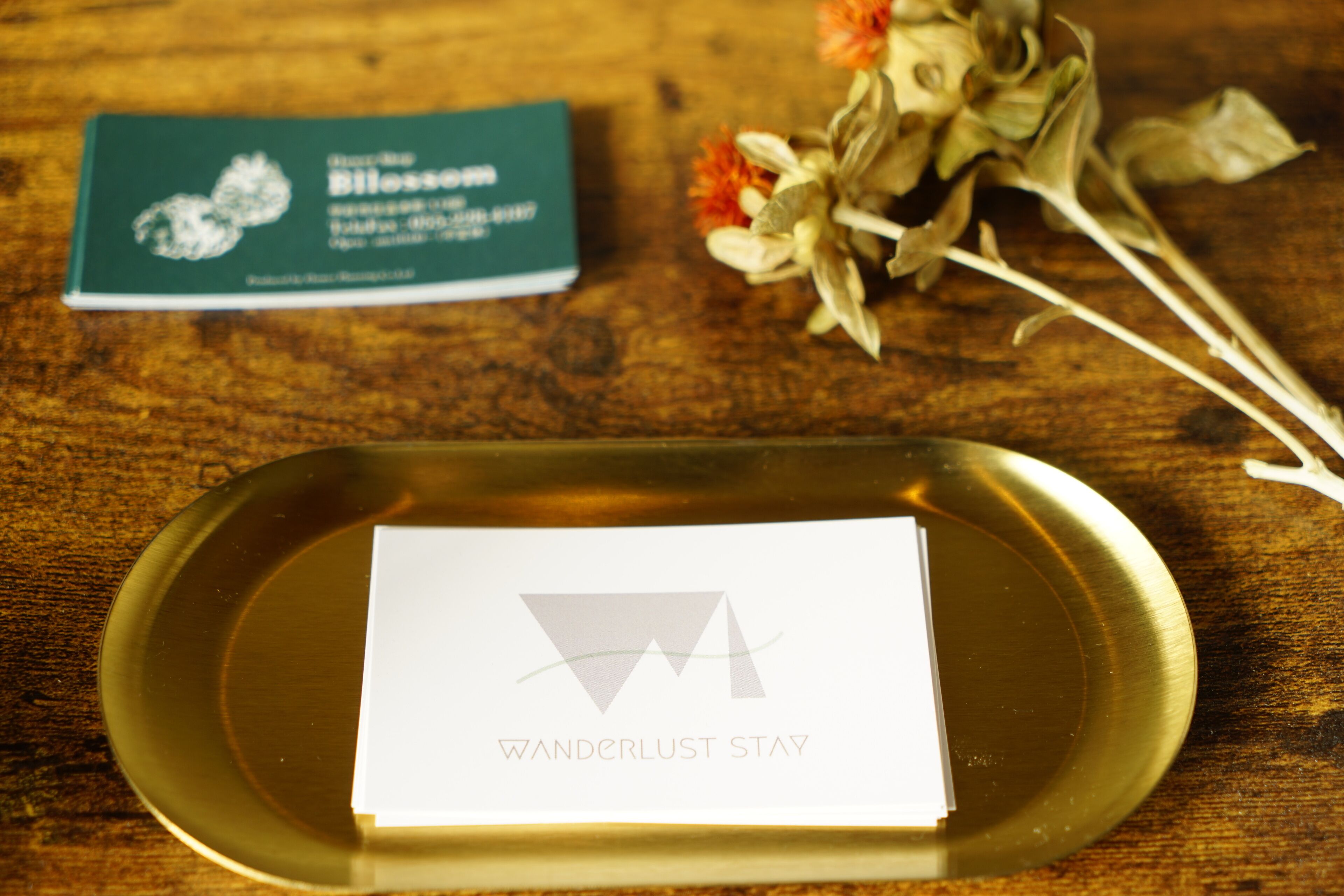 Una mesa de madera con un bandeja de vidrio que sostiene una tarjeta con la leyenda 'Wanderlust Stay' y una tarjeta de presentación con diseño floral.