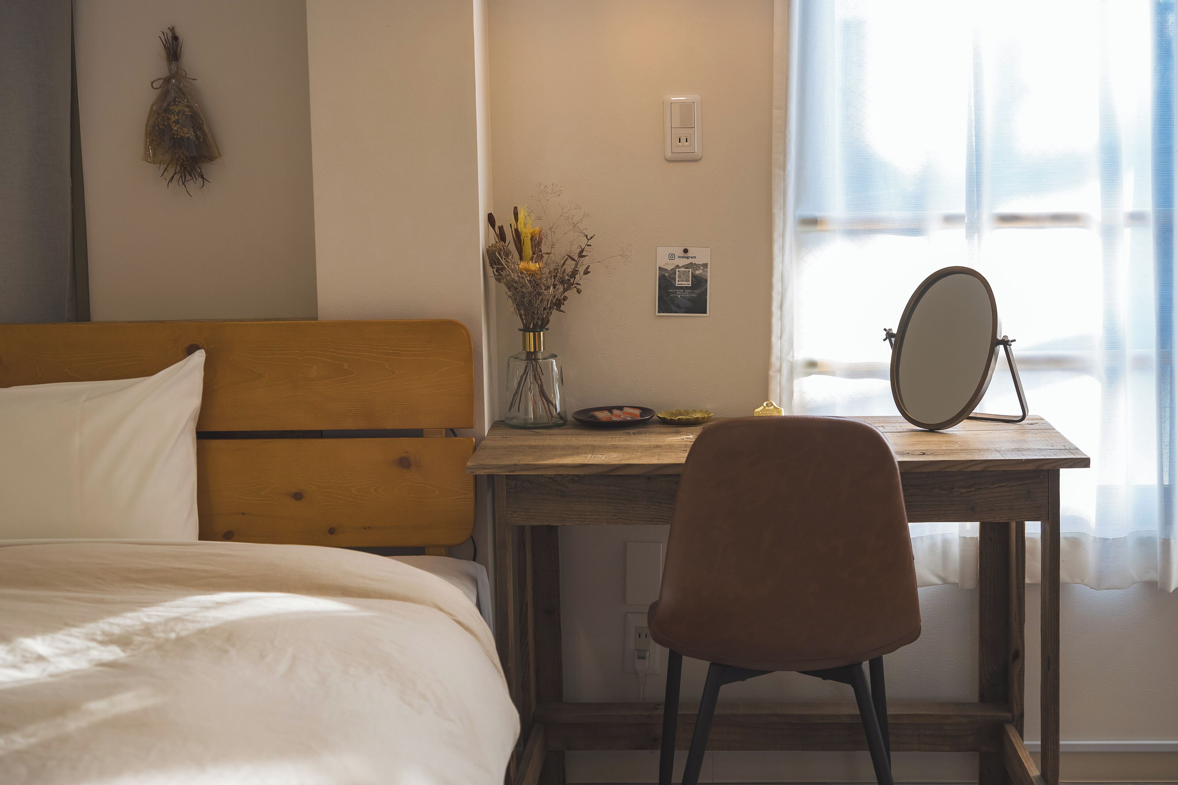 Quarto com cama de madeira, escrivaninha com cadeira, vaso com flores secas e espelho fixado na parede.