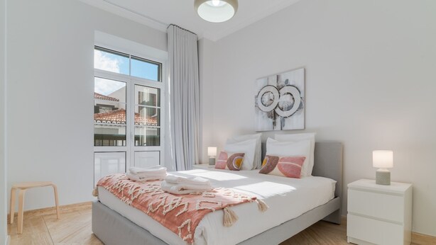 A modern bedroom with a large bed, a nightstand, a window with curtains, and a view of buildings outside. - Funchal City Nest II (Funchal)