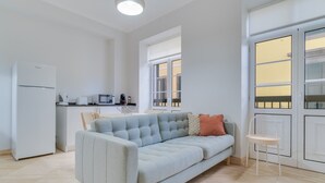 A modern living room with a light blue sofa, a small table, and a chair. There is a kitchen area with a refrigerator, microwave, and coffee machine. Large windows provide natural light and a view of the outside. - Funchal City Nest II (Funchal)