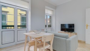 A modern living room with a grey sofa, a wooden dining table with chairs, a TV on a wooden stand, and large windows with shutters. - Funchal City Nest II (Funchal)