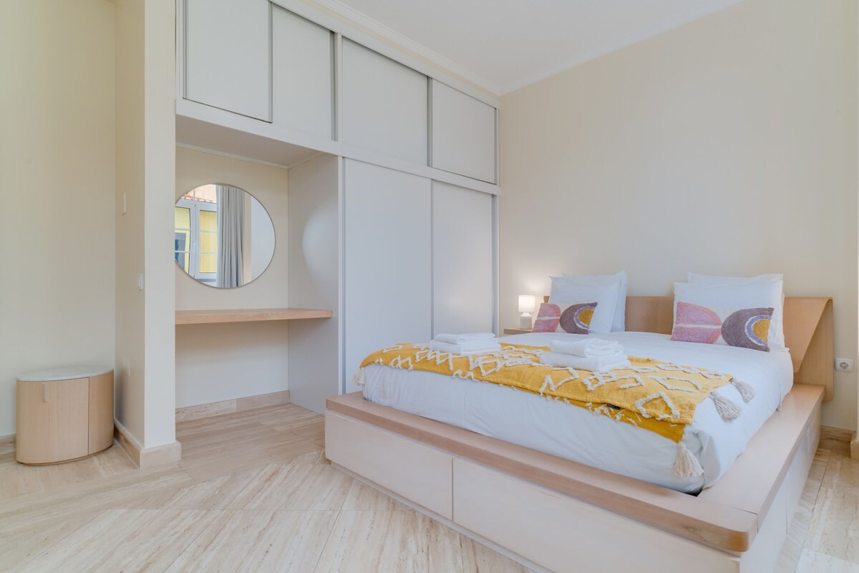 Un dormitorio moderno con cama, almacenamiento integrado, un espejo redondo y una mesita de noche.
