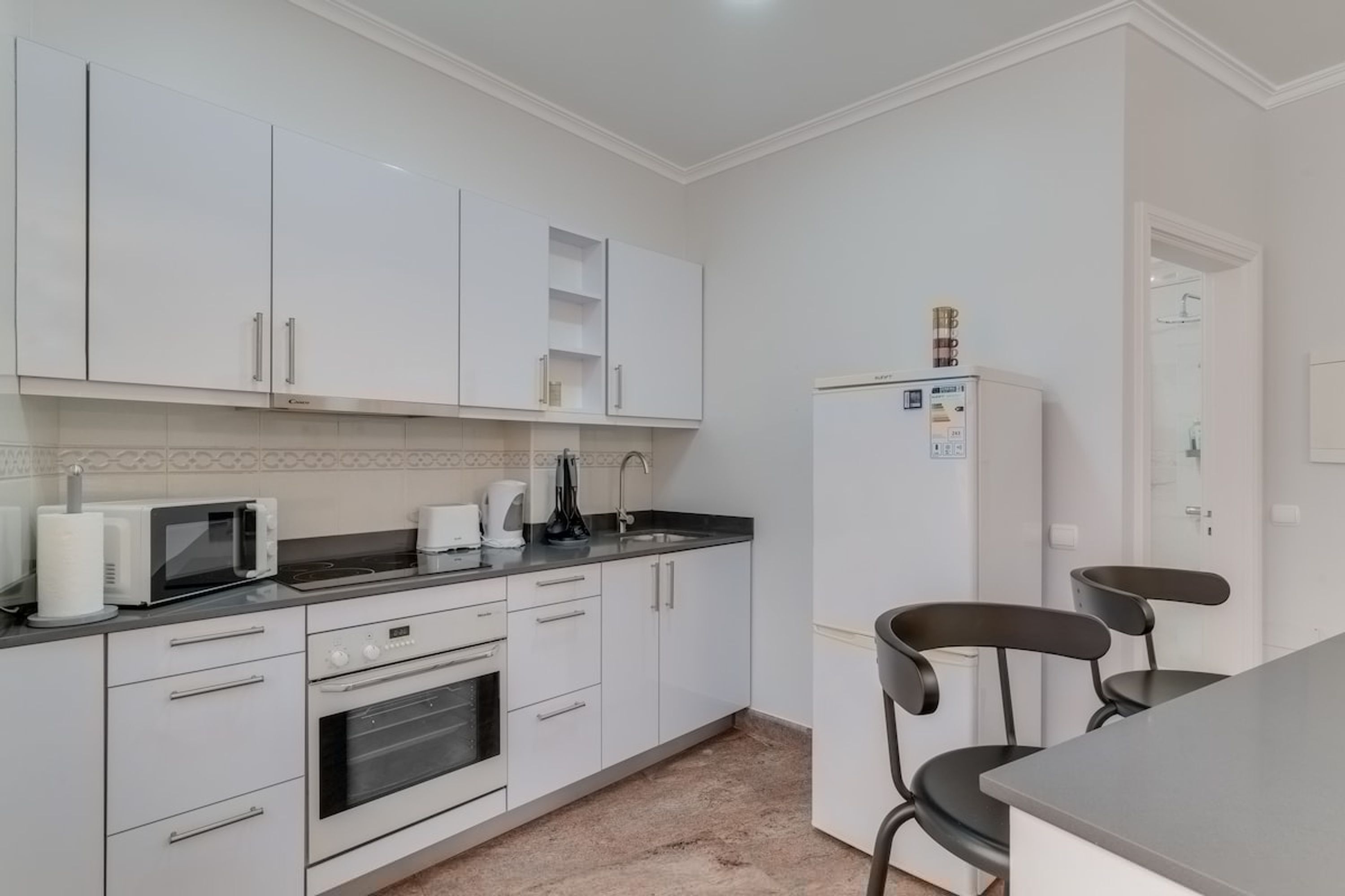 A modern kitchen with white cabinets, a microwave, a kettle, and a refrigerator. There are two black chairs at a table.