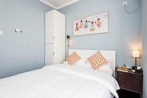 Un dormitorio con cama, ropa de cama blanca, almohadas con estampado naranja, una mesita de noche con lámpara y un cuadro colgado en la pared.