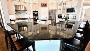A modern kitchen with white cabinets, stainless steel appliances, and a glass-top island with chairs. - Coastal Club Beach House w/ Resort Style Amenities (Lewes)