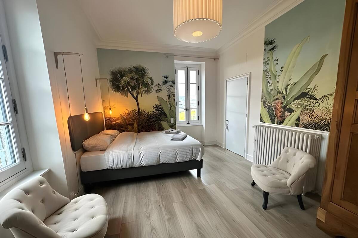 A bedroom with a bed, two chairs, a radiator, a window, and a wall mural.