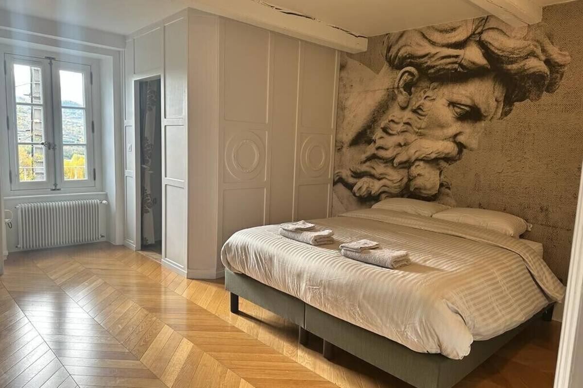 A bedroom with a large bed, a wooden floor, a window, and a wall with a textured mural.