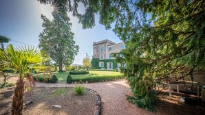 A well-maintained garden with a large tree, a palm, and a neatly trimmed lawn leading to a classical building with multiple windows and a stone facade.