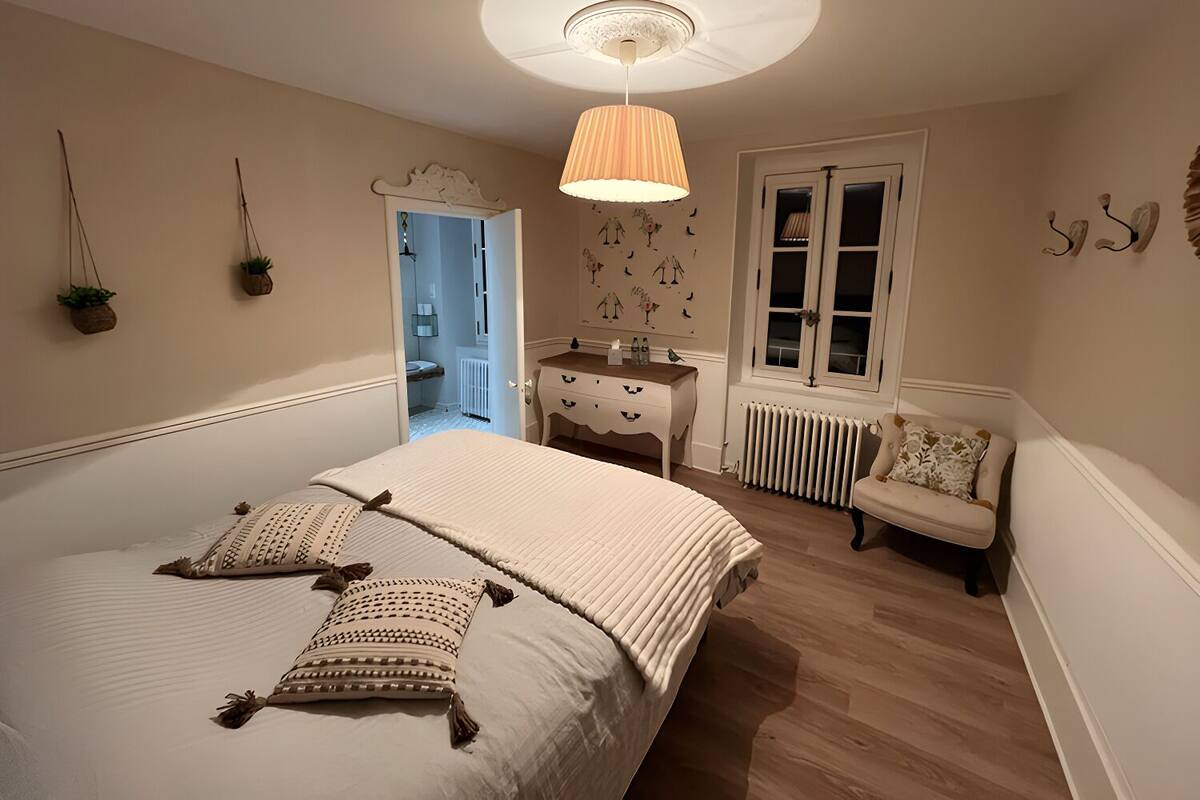 A bedroom with a large bed, a dresser, a chair, a radiator, and a door leading to another room.