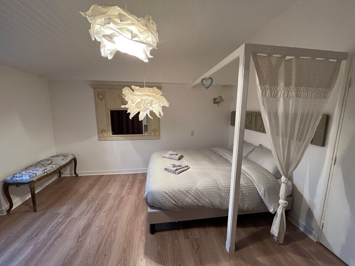 Een slaapkamer met hemelbed, een bankje, een spiegel en een wandlamp.