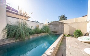 Ein rechteckiger Pool mit Steinrand, umgeben von Grünflächen und einem kleinen Garten mit einer Fontäne.
