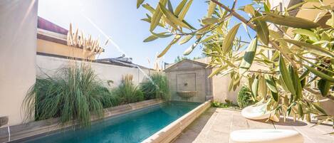Una piscina con borde de piedra, rodeada de vegetación y un pequeño edificio.