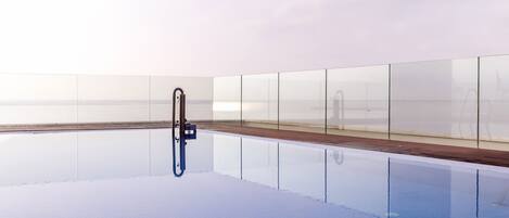 An empty swimming pool with a wooden deck and glass walls reflecting the sky.