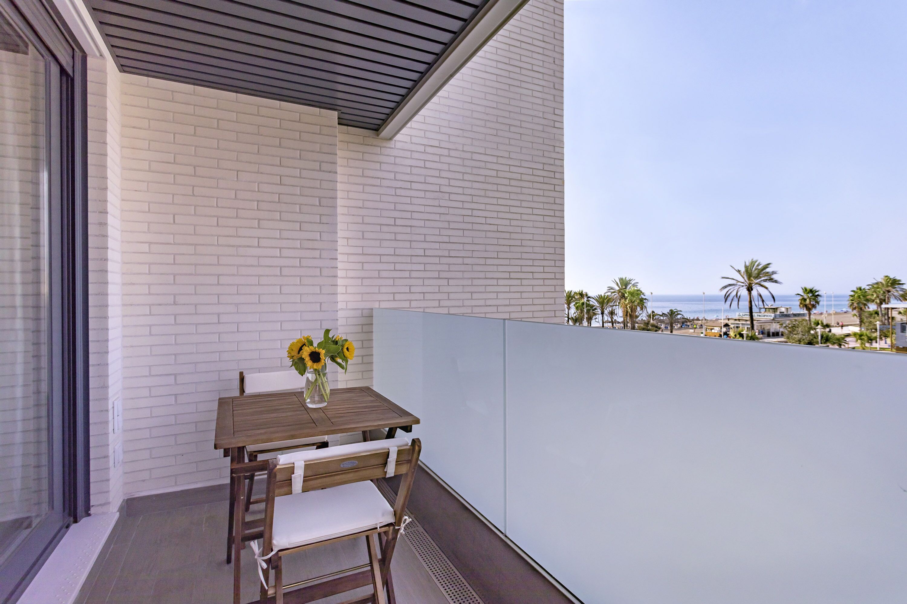 A balcony with a wooden table and chairs, a vase with sunflowers, and a view of the beach and palm trees.