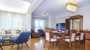 Een ruime woonkamer met een bank, blauwe fauteuils, een eettafel met stoelen, een houten kast en een televisie.