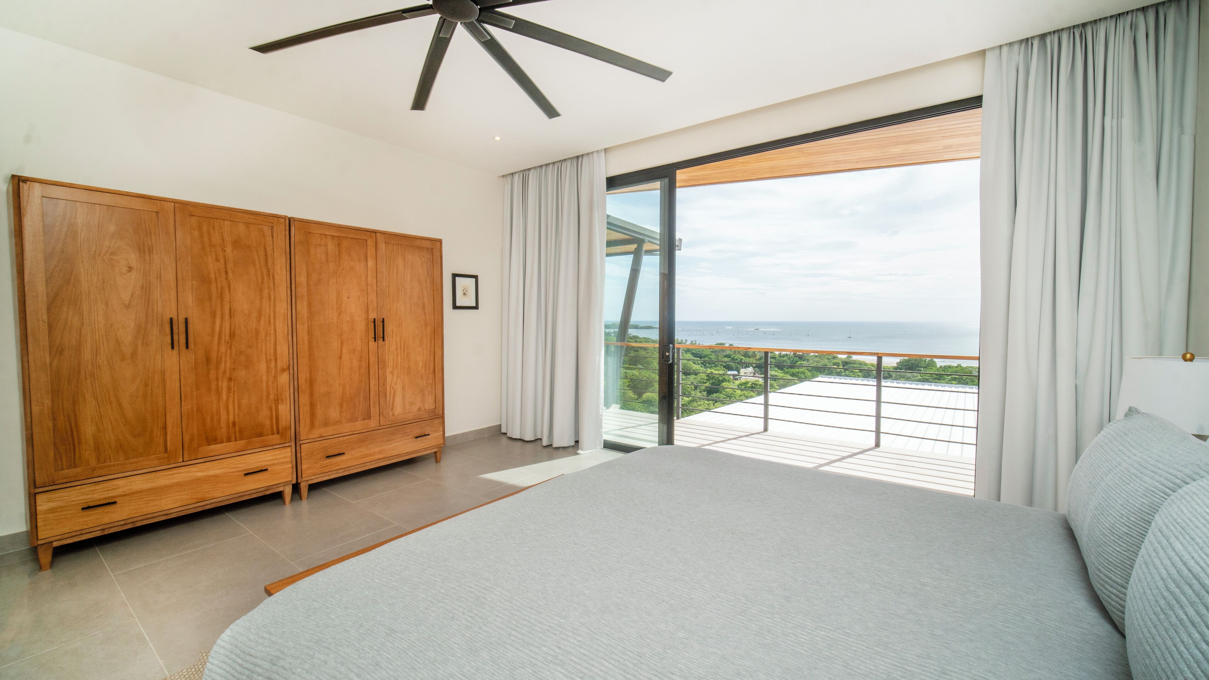 A bedroom with a large bed, a wooden wardrobe, a ceiling fan, and a balcony with a view of the sea.