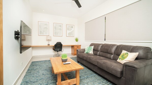 A modern living room with a brown leather sofa, a wooden coffee table, a black office chair, a small desk, and a mounted TV.