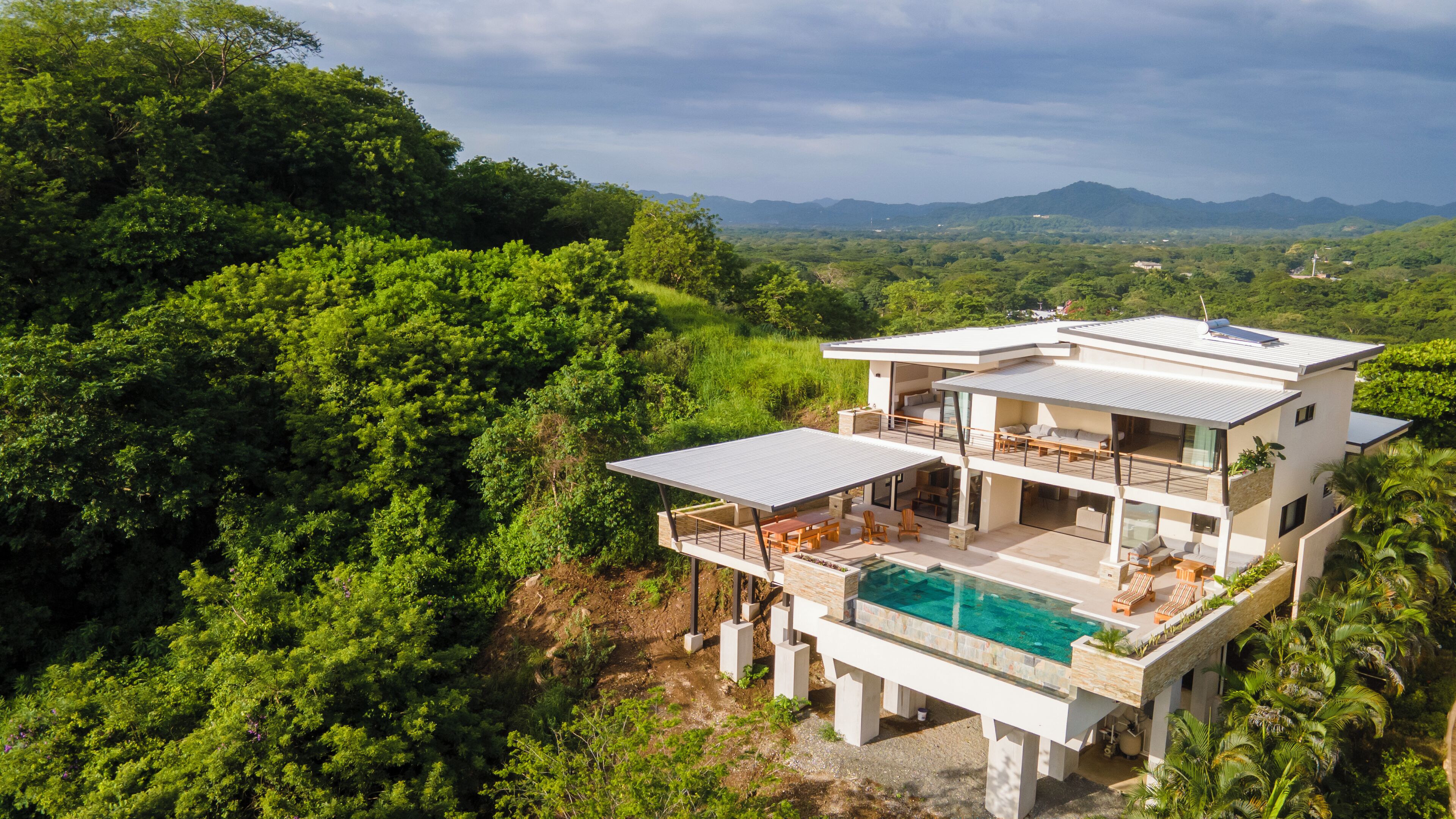 A modern two-story house with a pool, surrounded by lush greenery and mountains in the background.