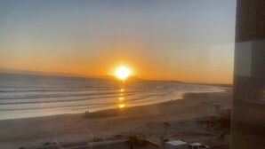Blick auf einen Strand bei Sonnenuntergang über dem Meer, einige Personen sind unterwegs und ein Gebäude im Vordergrund.