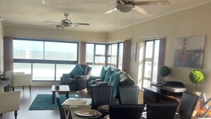 Ein modernes Wohnzimmer mit Essbereich, großen Fenstern, Deckenventilatoren und Blick aufs Meer.