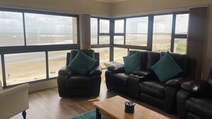 Ein Wohnzimmer mit zwei Ledersofas, einem Holztisch und Blick auf den Strand durch große Fenster.
